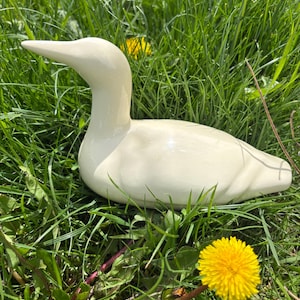 Può includere: Una statuetta di anatra in ceramica color crema con una finitura liscia e lucida. L'anatra è raffigurata in posizione di riposo con il collo teso verso l'alto e la testa leggermente girata di lato, mostrando una forma semplice e pulita.