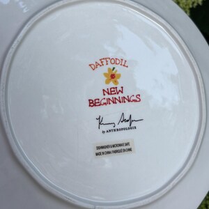 Może przedstawiać: Biały ceramiczny talerz z napisem "DAFFODIL NEW BEGINNINGS" w kolorze czerwonym i ż&oacute;łtym. Talerz ma kwiatowy wz&oacute;r i podpis. Talerz jest od Anthropologie.