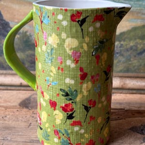 May include: A green ceramic pitcher with a floral pattern and a green handle. The pitcher has a white interior and is decorated with red, yellow, and blue flowers.
