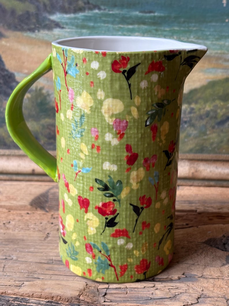 May include: A green ceramic pitcher with a floral pattern and a green handle. The pitcher has a white interior and is decorated with red, yellow, and blue flowers.