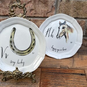 May include: Two white decorative plates with scalloped edges. One plate features a horseshoe with the text "very lucky". The other plate has a horse head and the text "happy you're here". Both plates are on a wooden surface.