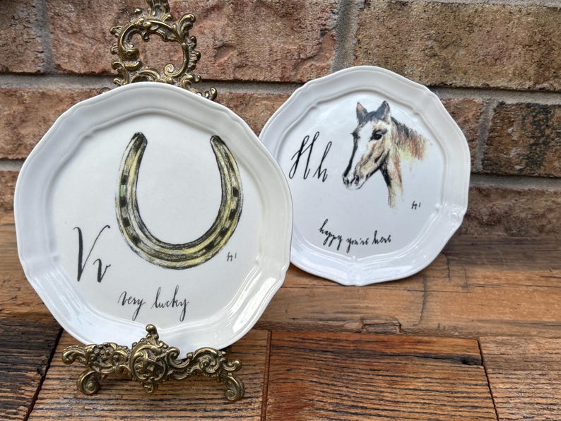 May include: Two white decorative plates with scalloped edges. One plate features a horseshoe with the text "very lucky". The other plate has a horse head and the text "happy you're here". Both plates are on a wooden surface.