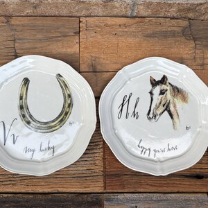 May include: Two white ceramic plates with scalloped edges. One plate features a horseshoe illustration with the text "very lucky." The other plate displays a horse head illustration with the text "happy you're here."