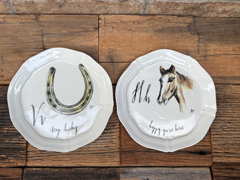 May include: Two white ceramic plates with scalloped edges. One plate features a horseshoe illustration with the text "very lucky." The other plate displays a horse head illustration with the text "happy you're here."