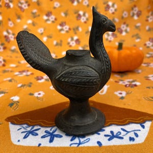 May include: A dark bronze-coloured bird-shaped candle holder with a long neck and a flared tail. The candle holder is on a round base. The background includes a pumpkin and a floral patterned fabric.