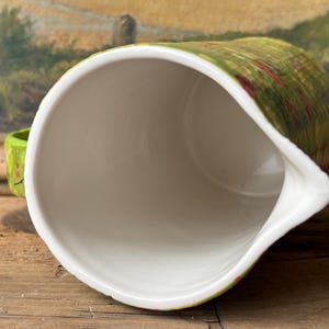 May include: A white ceramic pitcher with a green and red floral pattern. The pitcher has a wide mouth and a spout. The inside of the pitcher is white.