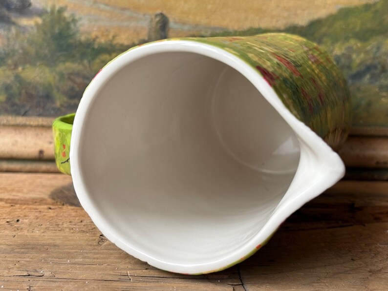 May include: A white ceramic pitcher with a green and red floral pattern. The pitcher has a wide mouth and a spout. The inside of the pitcher is white.