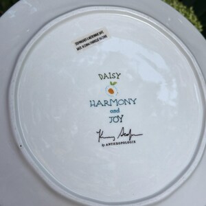 Może przedstawiać: Biały ceramiczny talerz z napisami "DAISY", "HARMONY and JOY" w niebieskim skrypcie. Talerz ma kwiatowy wz&oacute;r i podpis "K. Sedin" by Anthropologie. Można myć w zmywarce i używać w kuchence mikrofalowej.