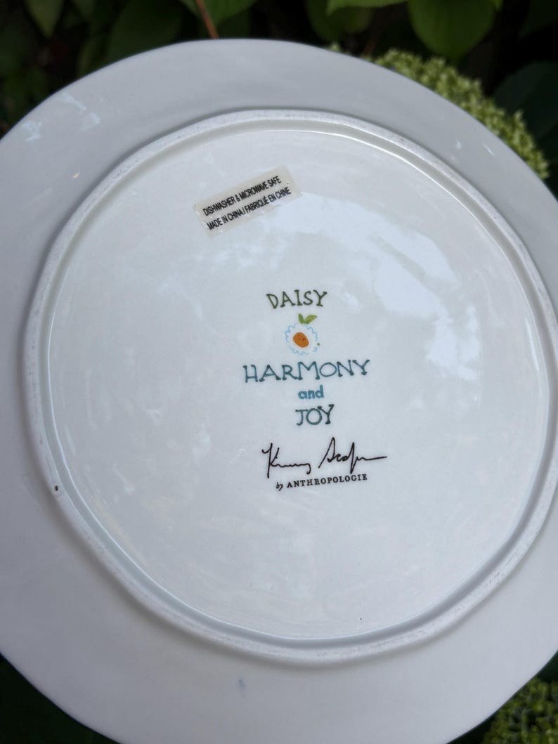 Może przedstawiać: Biały ceramiczny talerz z napisami "DAISY", "HARMONY and JOY" w niebieskim skrypcie. Talerz ma kwiatowy wz&oacute;r i podpis "K. Sedin" by Anthropologie. Można myć w zmywarce i używać w kuchence mikrofalowej.