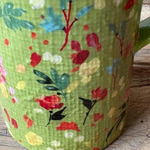May include: Green ceramic mug with a floral pattern in shades of red, yellow, blue, and green. The mug has a textured surface and a handle.