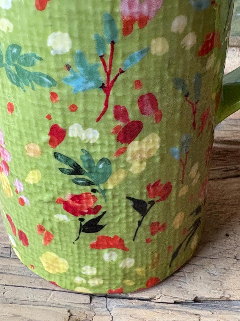 May include: Green ceramic mug with a floral pattern in shades of red, yellow, blue, and green. The mug has a textured surface and a handle.