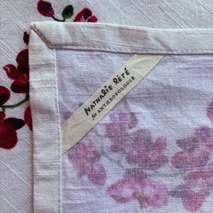 Puede incluir: Primer plano de un textil de lino blanco con un estampado floral en tonos rosas y morados. Se ve la esquina del textil, con un borde color crema. Una etiqueta dice "Nathalie R&eacute;t&eacute; para Anthropologie."