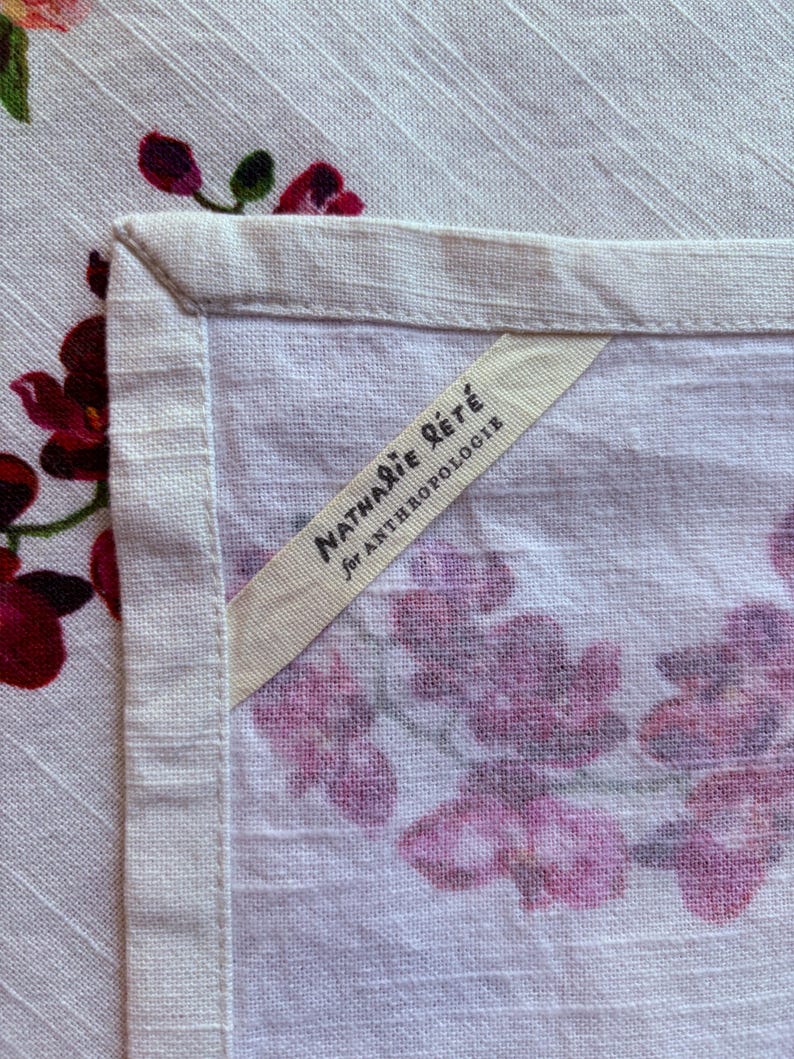 Puede incluir: Primer plano de un textil de lino blanco con un estampado floral en tonos rosas y morados. Se ve la esquina del textil, con un borde color crema. Una etiqueta dice "Nathalie R&eacute;t&eacute; para Anthropologie."