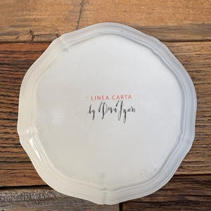 May include: A white, scalloped-edge ceramic plate with the text "LINEA CARTA by Dria Iyari" in black script and red font. The plate has a slightly irregular shape and a glossy finish. The background is a rustic wooden surface.
