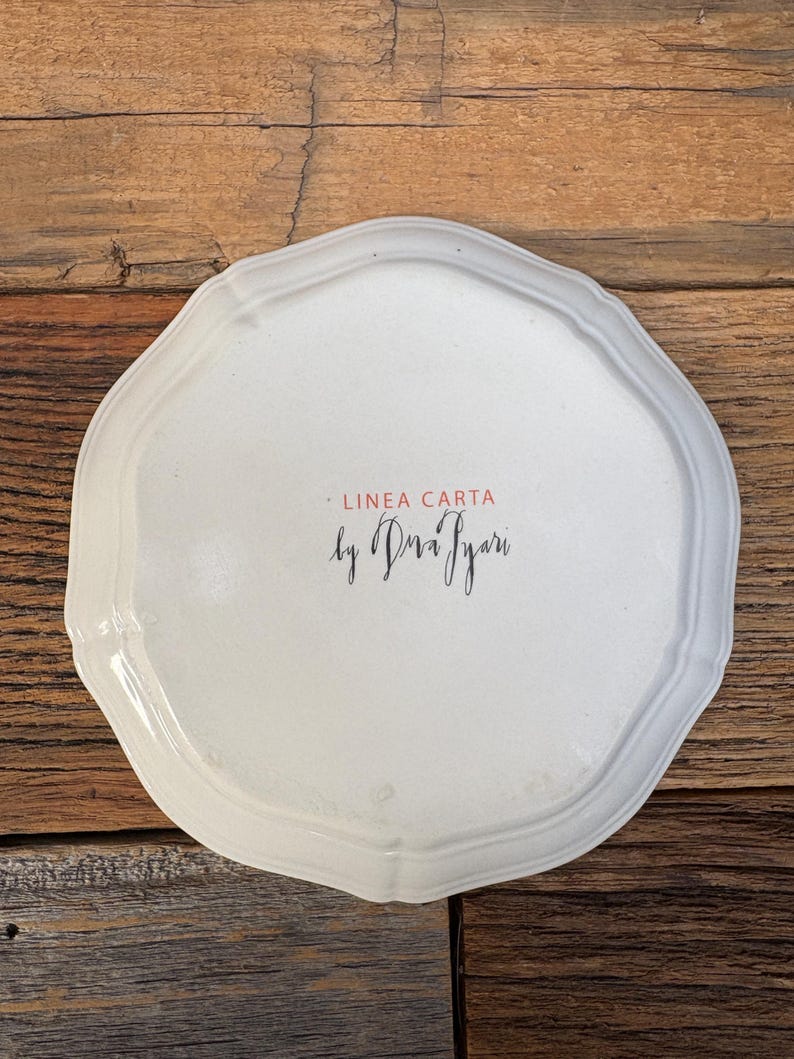 May include: A white, scalloped-edge ceramic plate with the text "LINEA CARTA by Dria Iyari" in black script and red font. The plate has a slightly irregular shape and a glossy finish. The background is a rustic wooden surface.