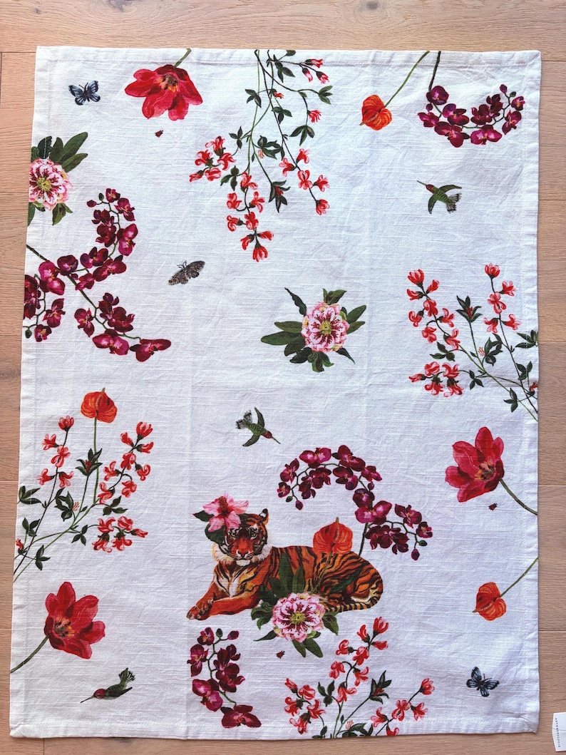 Puede incluir: Pa&ntilde;o de cocina rectangular blanco con un estampado floral y animal vibrante. El dise&ntilde;o presenta tulipanes rojos, orqu&iacute;deas, mariposas, colibr&iacute;es y un tigre. El tigre est&aacute; adornado con flores. El pa&ntilde;o est&aacute; hecho de un material suave y absorbente.