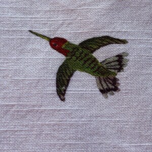Puede incluir: Bordado detallado de un colibr&iacute; en vuelo, con cabeza roja, cuerpo verde y alas oscuras. El ave se encuentra sobre un fondo de tela beige claro y texturizado, mostrando los colores vibrantes y el bordado.