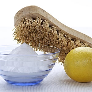 May include: A white bowl filled with baking soda, a yellow lemon, and a wooden brush with natural bristles.