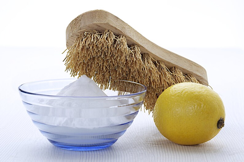 May include: A white bowl filled with baking soda, a yellow lemon, and a wooden brush with natural bristles.