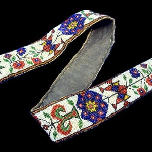 May include: A beaded belt with a white background and colourful floral and geometric patterns. The belt features red, blue, green, and yellow beads. The belt has a grey leather backing.