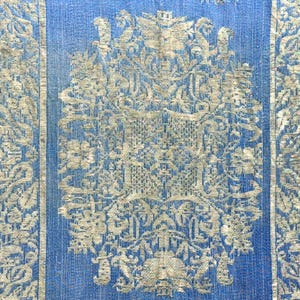May include: Close-up of a blue textile with a detailed, ornate design. The central panel features a large, symmetrical pattern with floral and architectural elements in a lighter color. Vertical stripes frame the central design.