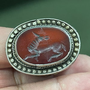 Puede incluir: Un anillo de plata con una piedra ovalada de cornalina que presenta una imagen tallada de un ciervo corriendo. La piedra está engastada en un engaste de plata con un borde decorativo de pequeñas cuentas.