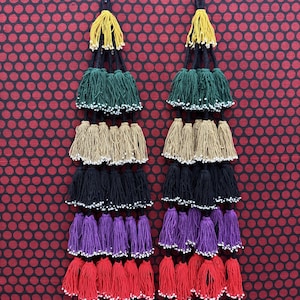 Puede incluir: Un par de pendientes largos de borlas escalonadas con cinco filas horizontales de borlas de hilo en amarillo, verde, beige, negro, morado y rojo. Cada borla está rematada con pequeñas cuentas blancas en la parte inferior, suspendidas de un cordón negro.