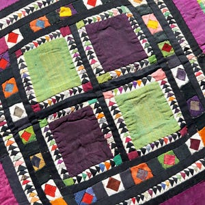 May include: Close-up of a colorful, handmade quilt with a patchwork design. The quilt features squares of green and purple fabric, bordered by black fabric and white triangles. The edges are trimmed with orange and purple fabric.