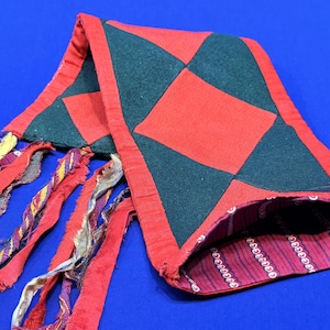 Puede incluir: Un artículo de tela roja y verde con un patrón de diamantes. El artículo tiene un borde rojo y está forrado con una tela estampada. Tiras de tela roja con acentos coloridos cuelgan del artículo.