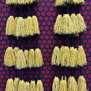 May include: A decorative wall hanging featuring two vertical rows of mustard yellow yarn tassels suspended from dark navy blue cords, set against a deep burgundy fabric background patterned with a repeating grid of dark purple polka dots.
