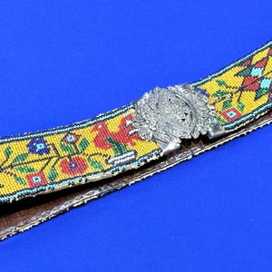 May include: A beaded belt with a yellow background and colourful floral and geometric patterns. The belt has a silver-coloured metal buckle. The belt is approximately 5 cm wide.