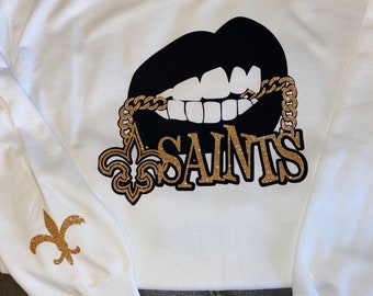 May include: White sweatshirt with a black and gold glitter design of a pair of lips with a gold chain and the word "SAINTS" on the front. The left sleeve has a gold glitter fleur-de-lis design.