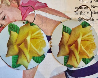 May include: A pair of circular earrings with a yellow rose and green leaves on a white backdrop. The earrings have silver-coloured hooks. The earrings are vintage in style.