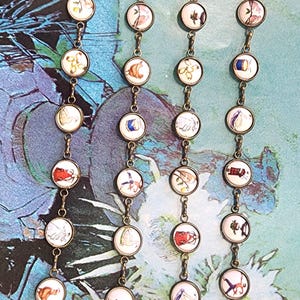 May include: Antique brass-toned eyeglass chains with round, illustrated charms. Each charm features a unique, colorful design. The chains have lobster claw clasps for easy attachment to glasses. The background is a watercolor-style painting.