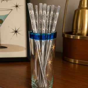 May include: A set of silver-toned metal cocktail stirrers with embossed figure designs, housed in a clear glass tumbler featuring a blue rim and gold starburst patterns. The stirrers have a textured, fan-shaped top and a slender, tapered body.