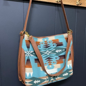 May include: A blue and brown patterned tote bag with a brown leather strap and a gold buckle. The bag is hanging on a wooden rack.