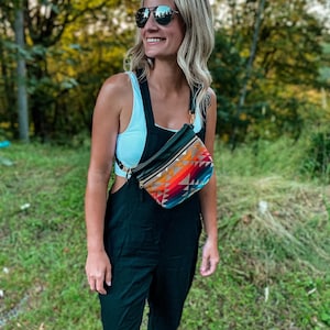 May include: A woman wearing a white tank top and black overalls with a colorful patterned fanny pack worn across her body. She is wearing sunglasses and smiling.