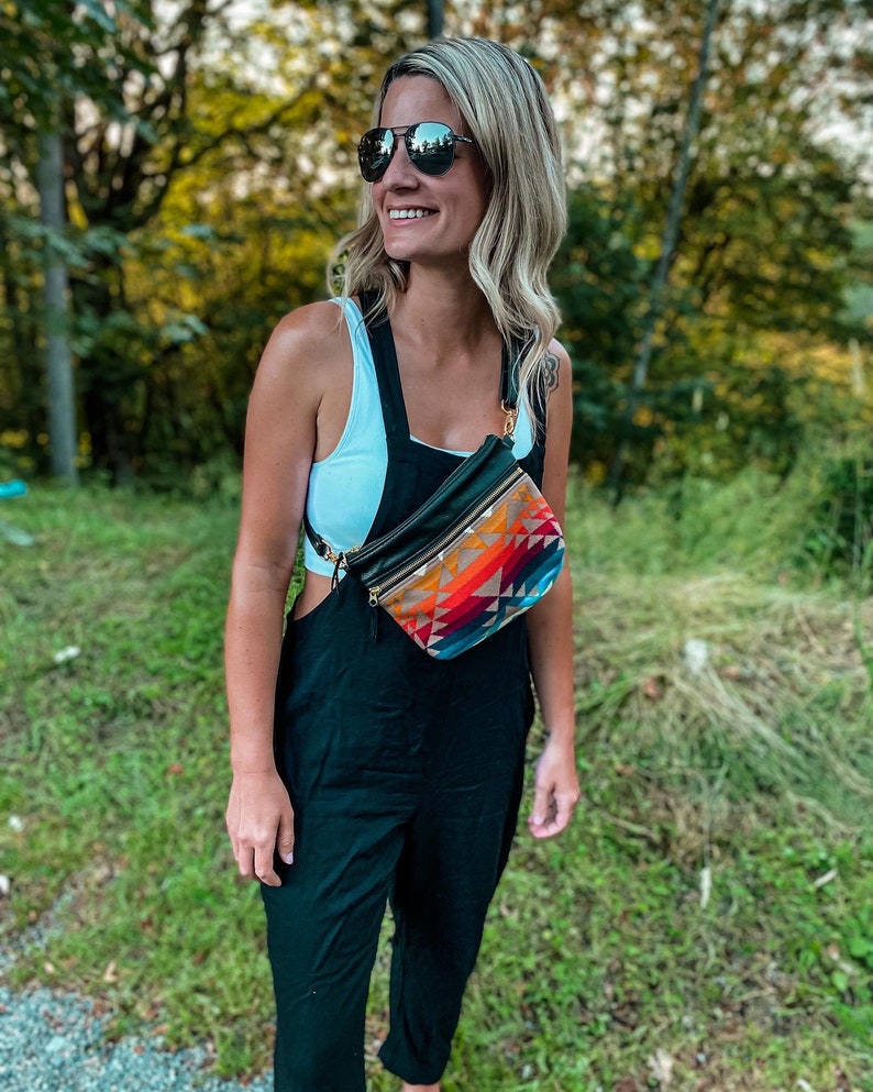 May include: A woman wearing a white tank top and black overalls with a colorful patterned fanny pack worn across her body. She is wearing sunglasses and smiling.