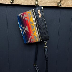 May include: A black leather crossbody bag with a colorful patterned flap. The flap has a geometric design with shades of blue, red, orange, yellow, and brown. The bag has a gold zipper and a leather strap with a gold buckle. The bag has a leather tag with the word "SALAITO" printed on it.