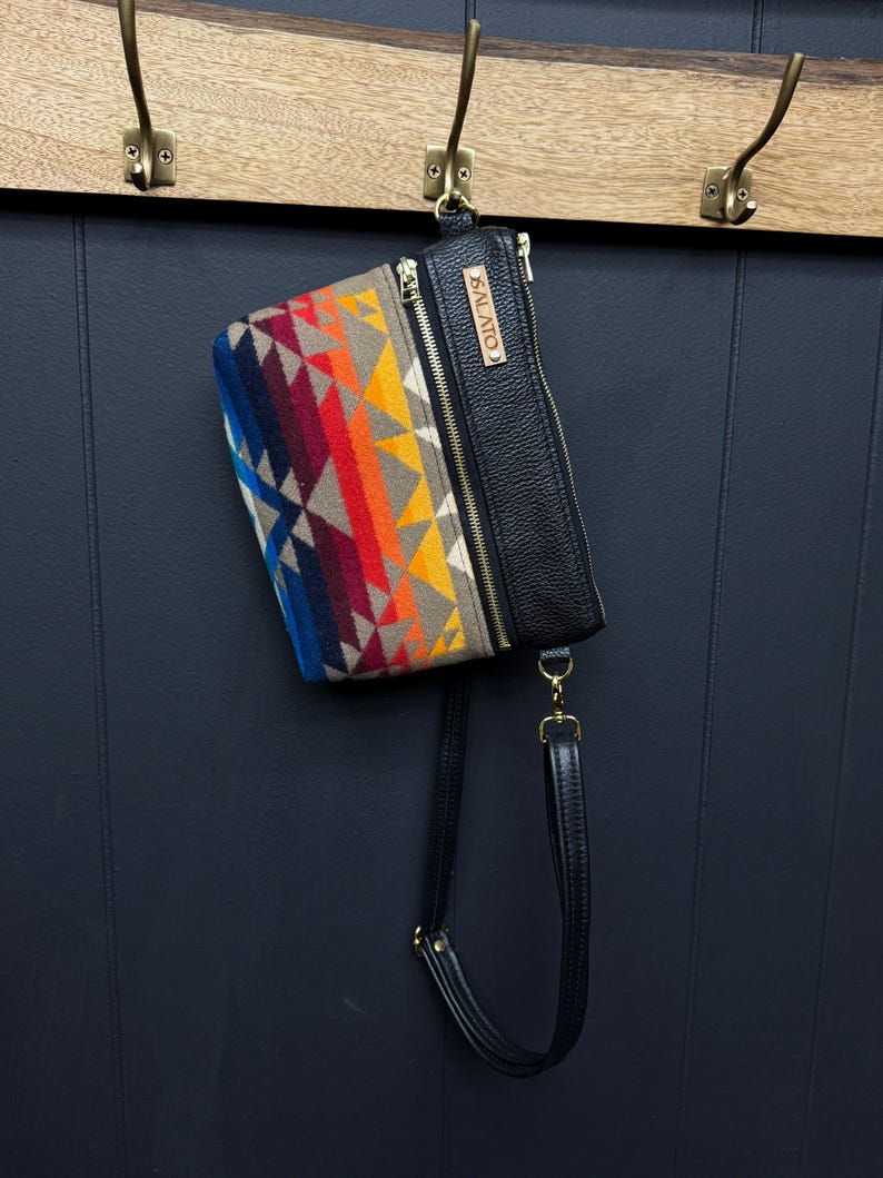 May include: A black leather crossbody bag with a colorful patterned flap. The flap has a geometric design with shades of blue, red, orange, yellow, and brown. The bag has a gold zipper and a leather strap with a gold buckle. The bag has a leather tag with the word "SALAITO" printed on it.