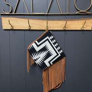 May include: A black and white patterned clutch with tan fringe hangs from a brass hook. The clutch has a geometric design and a tan leather fringe. The wall behind the clutch is dark gray.