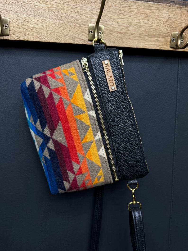 May include: A black leather crossbody bag with a colorful geometric patterned flap and a gold zipper. The bag has a brown leather tag with the text "SALATO" on it.