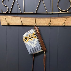 Peut inclure: Un sac bandoulière à motifs géométriques gris, jaunes et blancs est suspendu à un porte-manteau en bois avec des crochets en laiton. Le sac a un dessus et une bandoulière en cuir marron. Le mot "SALATO" est affiché au-dessus.
