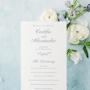 May include: A wedding ceremony program with a blue and white floral design. The program lists the order of events for the wedding of Caitlin and Alexander on September 28, 2024 at Swaka Riva Ranch.