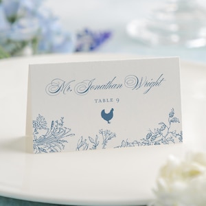 May include: A white place card with blue floral and chicken illustrations. The card reads "Mr. Jonathan Wright" and "Table 9" in elegant script. The card is set on a white plate, with blue and white flowers in the background.