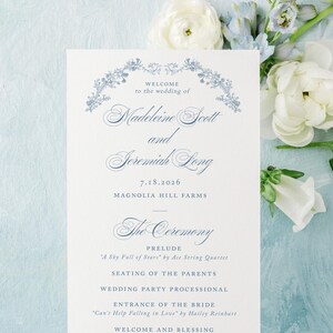 May include: A wedding program with elegant blue floral accents and text. The program lists the wedding details, including the names Madeleine Scott and Jeremiah Long, and the ceremony schedule. White and blue flowers surround the program.