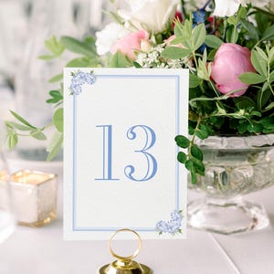 May include: A white table number card with a blue border and blue floral accents. The number 13 is printed in blue on the card. The card is held in a gold metal stand.