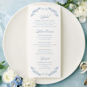 May include: A white menu card with a floral design and elegant script is placed on a white plate. The menu lists salad, main course, and dessert options. A vintage fork and spoon are tied with a ribbon, and flowers surround the plate.
