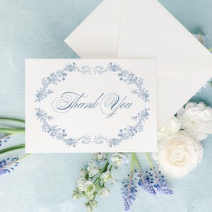 May include: A white card with the words "Thank You" in elegant script, surrounded by a blue floral wreath. An envelope and flowers, including white roses and bluebells, are arranged on a light blue surface.