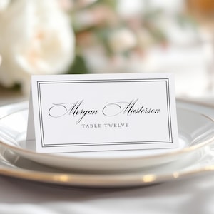 May include: White place card with a black border and the text "Morgan Masterson" and "Table Twelve" in black script font.